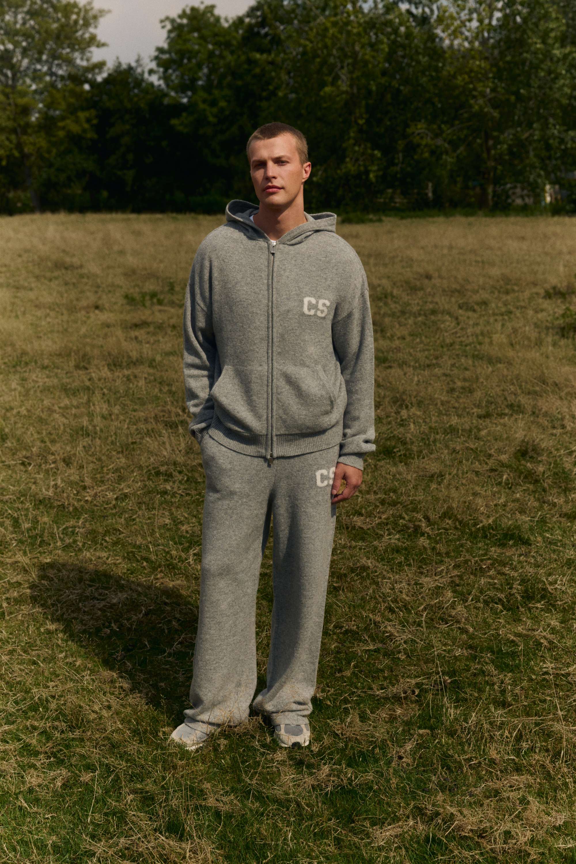 Man wearing a grey melange CASPARA woolknit zip jacket and matching knit pants in a grassy field.